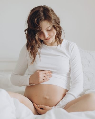 Happy pregnant woman in white holding belly bump and relaxing on white bed at home. Stylish pregnant mom hugging belly with love and care, waiting for baby. Motherhood concept ,maternity time
